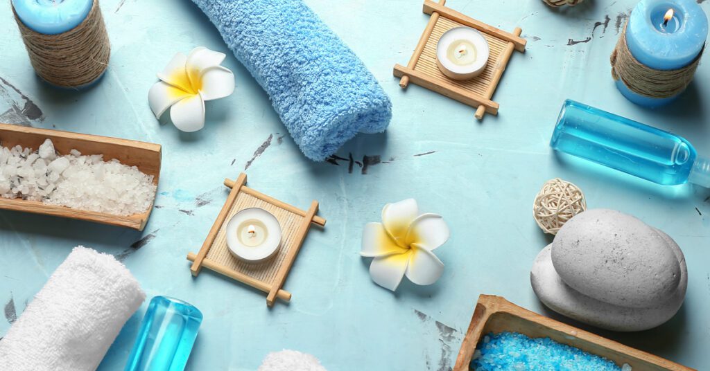An arrangement of spa accessories—rolled up towels, bath salts, candles, and essential oils—are against a blue background.