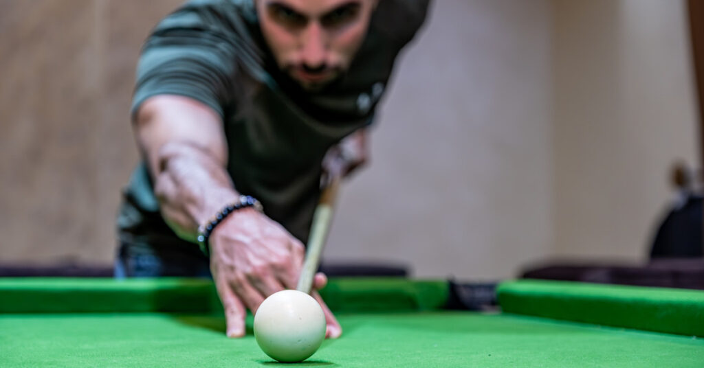 Male young adult with a beard standing behind a billiards table holding a cue stick aiming to hit the white ball.