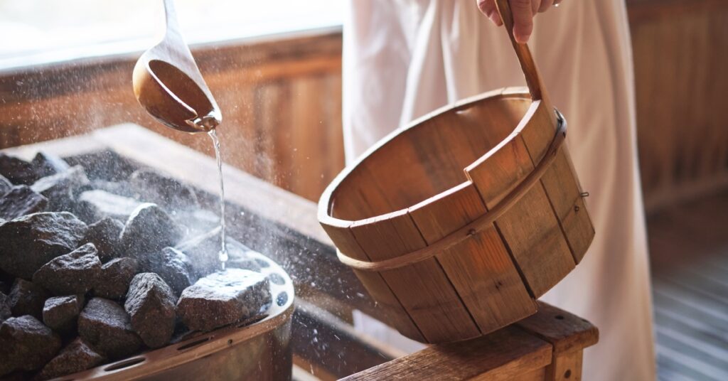 The bottom half of a man in a towel taking water from a bucket. He is pouring the water onto hot stones in a wooden sauna.