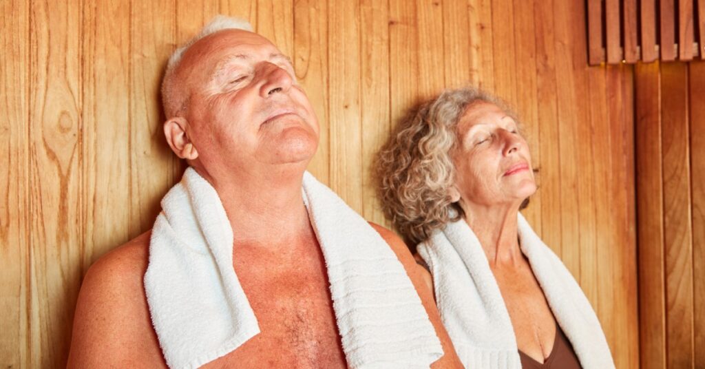 Male and female seniors sitting next to each other inside a wooden sauna wearing small towels around their necks while relaxing.