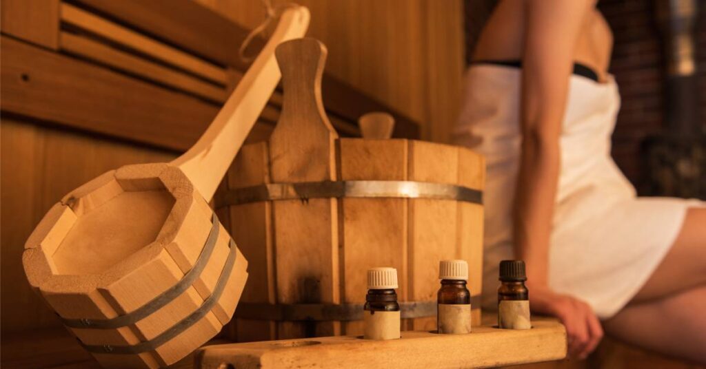 Wooden accessories inside a sauna with aromatic oils on top of a wooden board with a woman wrapped in a towel behind.