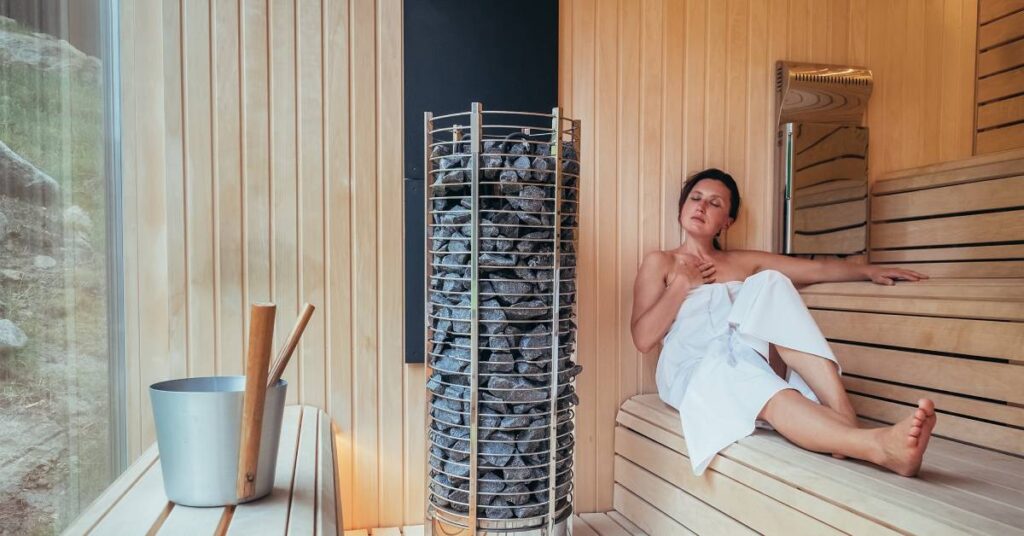 A woman wrapped in a big white towel is sitting inside a sauna with a metal bucket, a stone tower, and a large window.