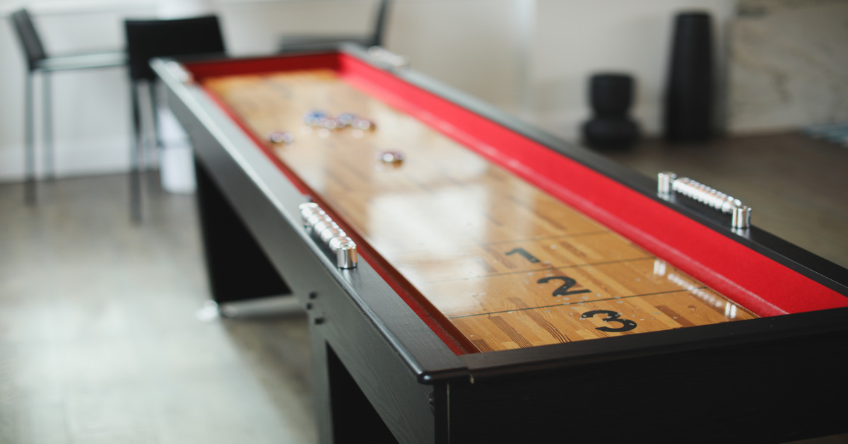 Shuffleboard table with a black finish, a red trim, and a wooden playing surface inside a room with tall chairs.