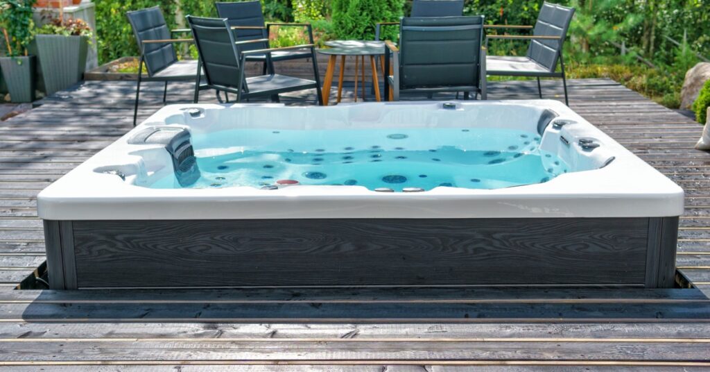 A large swim spa placed on a wooden deck holds bright blue water. Six chairs are nearby gathered around a table.