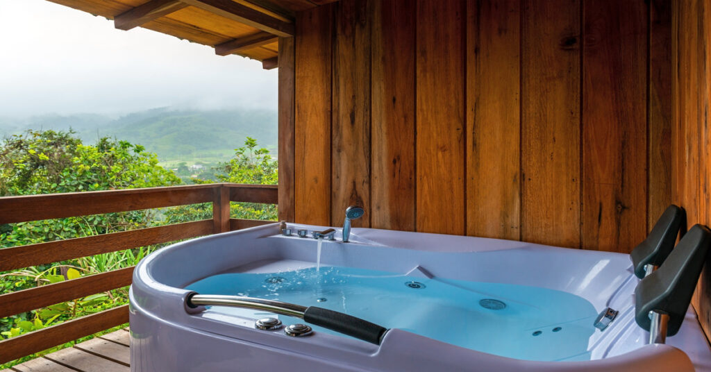 A hot tub in a shaded area on a wooden balcony overlooking trees and lush, green forest hills in the distance.