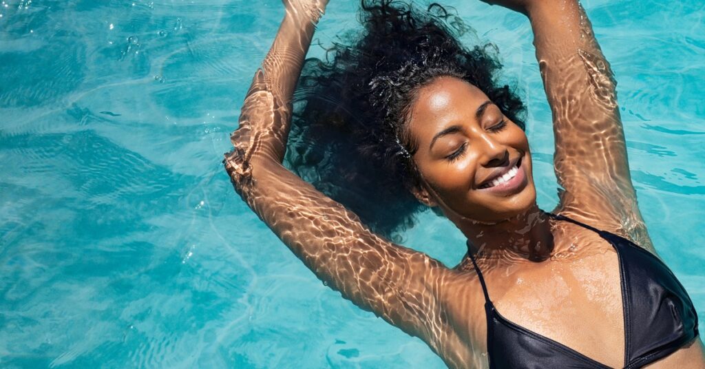 A woman wearing a black swimsuit is smiling and floating on her back in a pool. The water is bright blue.