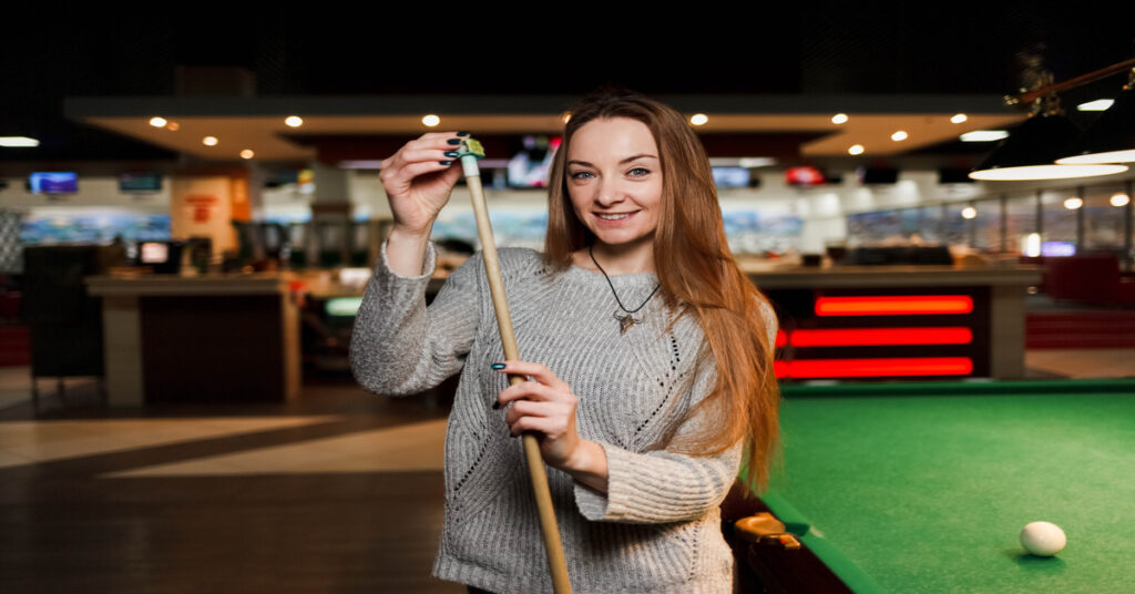 A smiling woman with long hair chalking her cue stick beside a pool table with a cue ball on the felt.