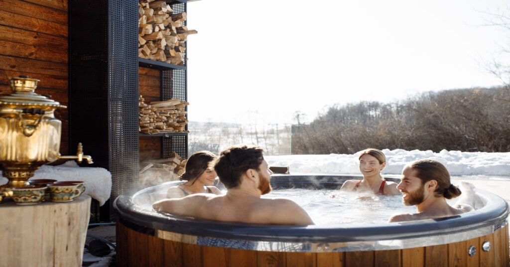 A smiling, mixed-gender group sitting in a steaming hot tub near a snow-covered expanse lined with trees.