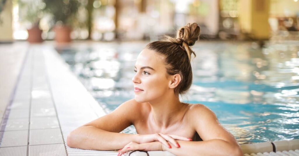 A young blonde woman with brightly painted nails resting her arms on the tiled edge of a large indoor pool.