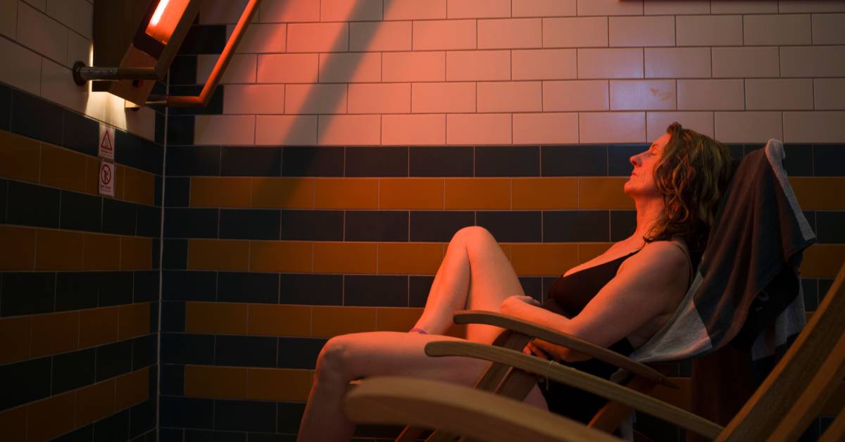 A middle-aged woman in a bathing suit sleeping on a wooden chair in front of a large infrared sauna light.