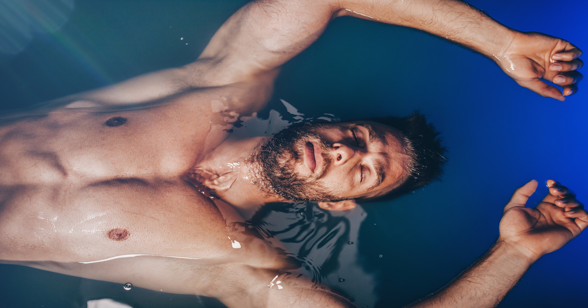 A bearded man, with his eyes closed, floats on his back in a pool of deep-blue water.