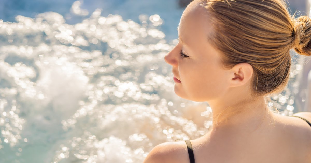 A young blonde woman in a black bathing suit sitting inside a bubbling hot tub with her eyes closed.