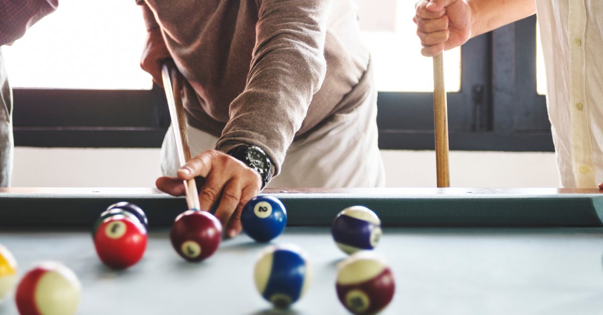 Two men play billiards in a home. One is taking a shot with his cue, while the other stands on the right.