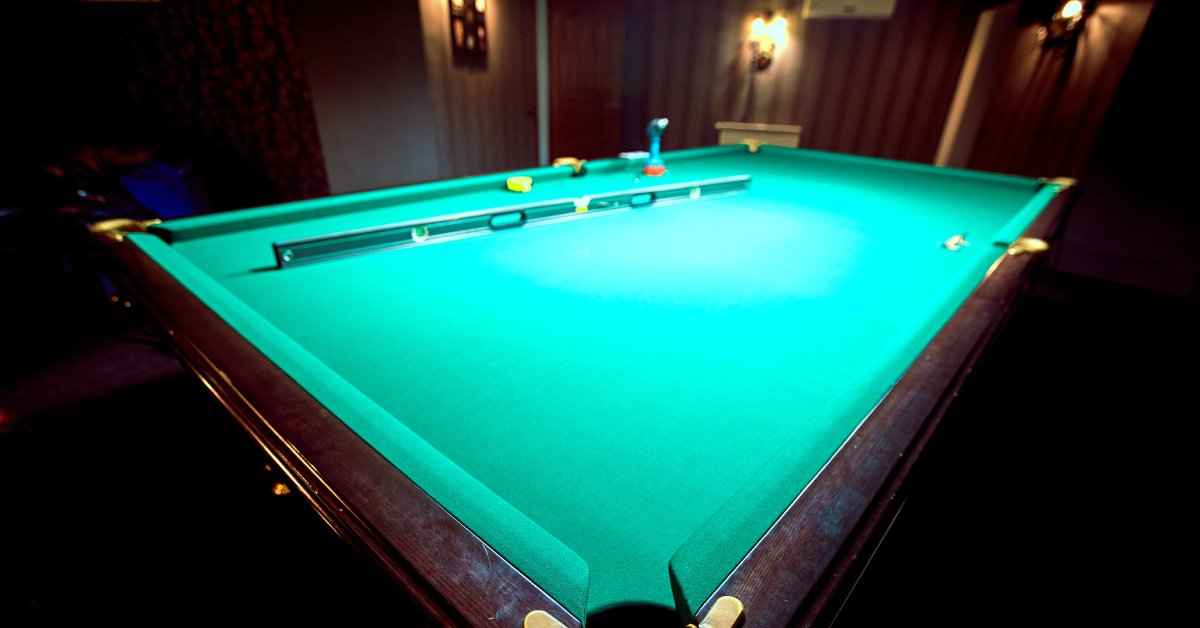 A carpenter's level, drill, and measuring tape sit on a billiards table with green felt, located in a room with wood paneling.
