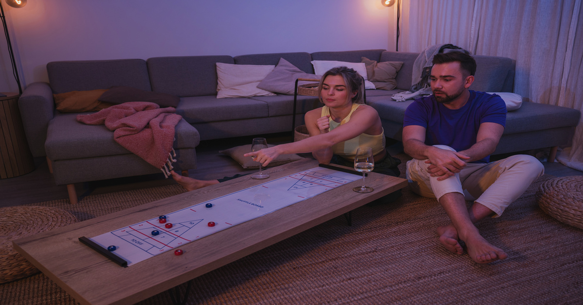 A young man and woman sit on the floor of their living room in front of a shuffleboard table. The woman points at the table.