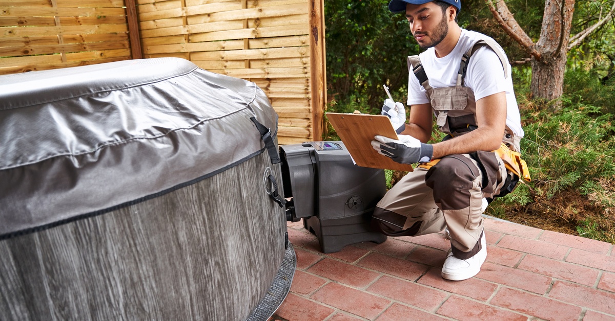 A male technician wearing brown overalls and a blue hat crouches in front of a covered hot tub, making notes on a clipboard.