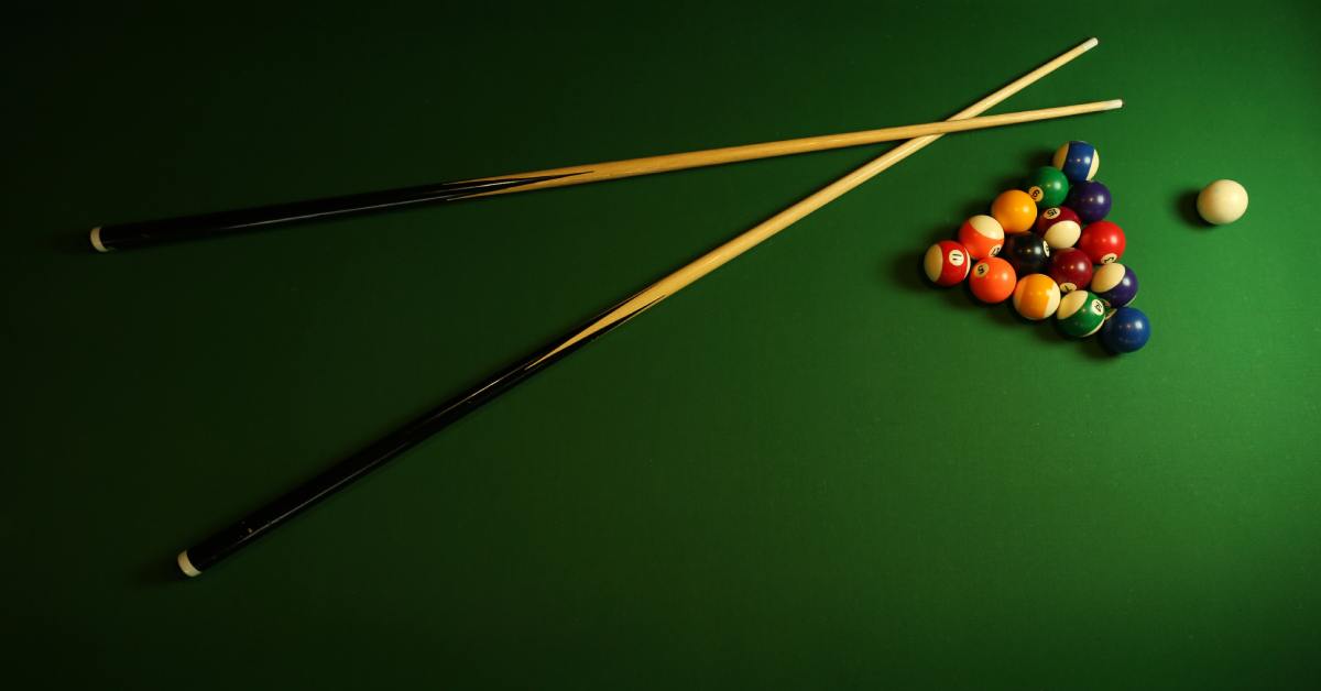 A pair of pool cues and a full set of pool balls rest on the green felt of a pool table.