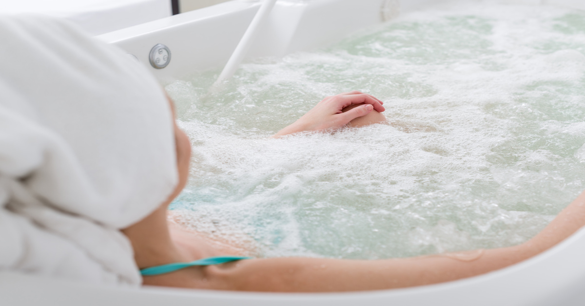 A woman with a white towel wrapped around her head relaxes in a small spa, water bubbling around her.