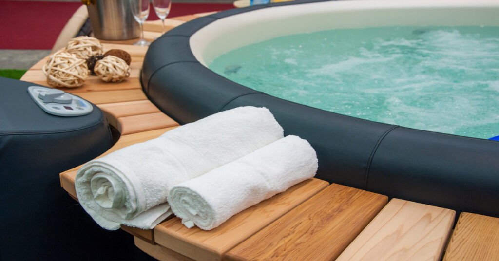 A circular hot tub with a black rubber rim and a wooden deck surrounding the rim. Two white towels and various accessories sit on the deck.