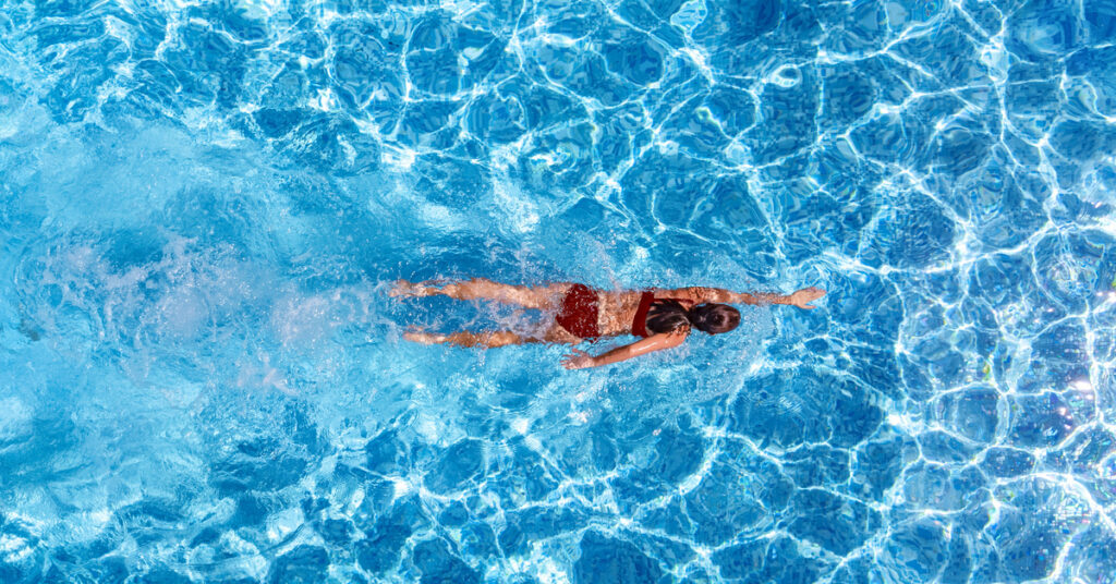 Overhead view of a woman in a red bathing suit swimming in an expanse of turquoise pool water.