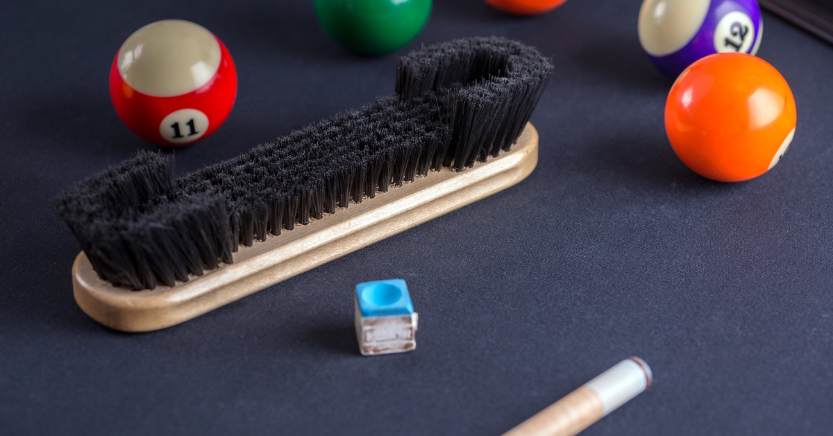 A felt brush lying atop a gray-felted pool table, surrounded by five pool balls, a blue chalk square, and a pool cue.