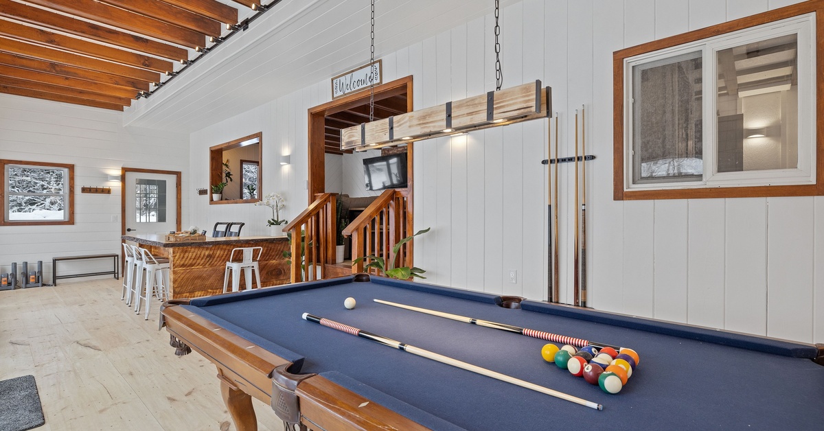 A gray-felted pool table sitting in the middle of an upscale game room with wooden decorative elements and a bar.