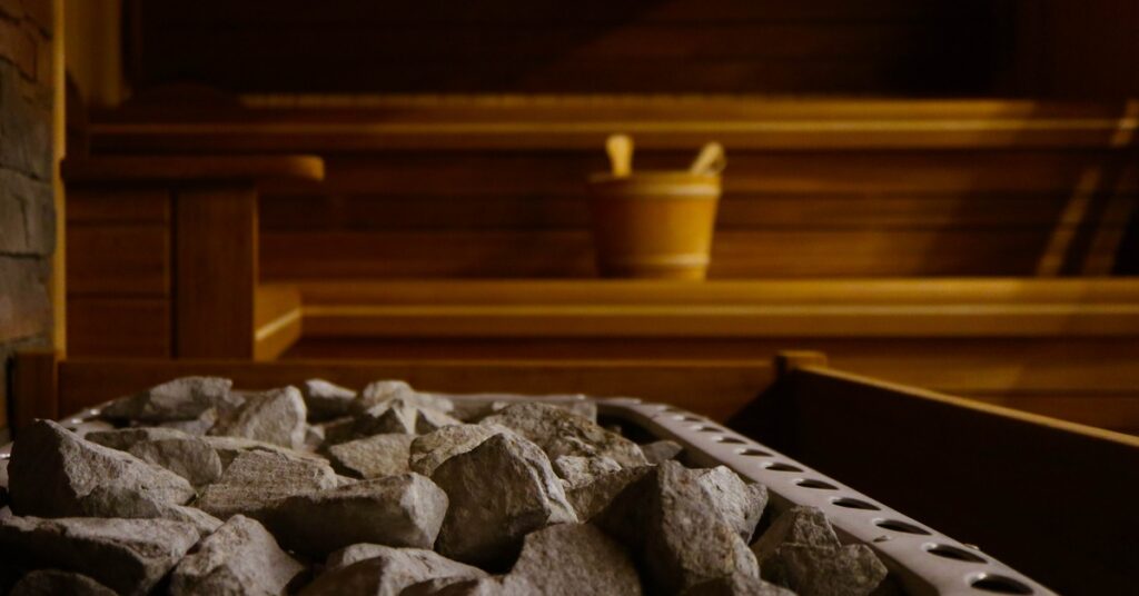 A pile of sauna stones in the foreground, with a wooden sauna interior and a wooden bucket in the background.