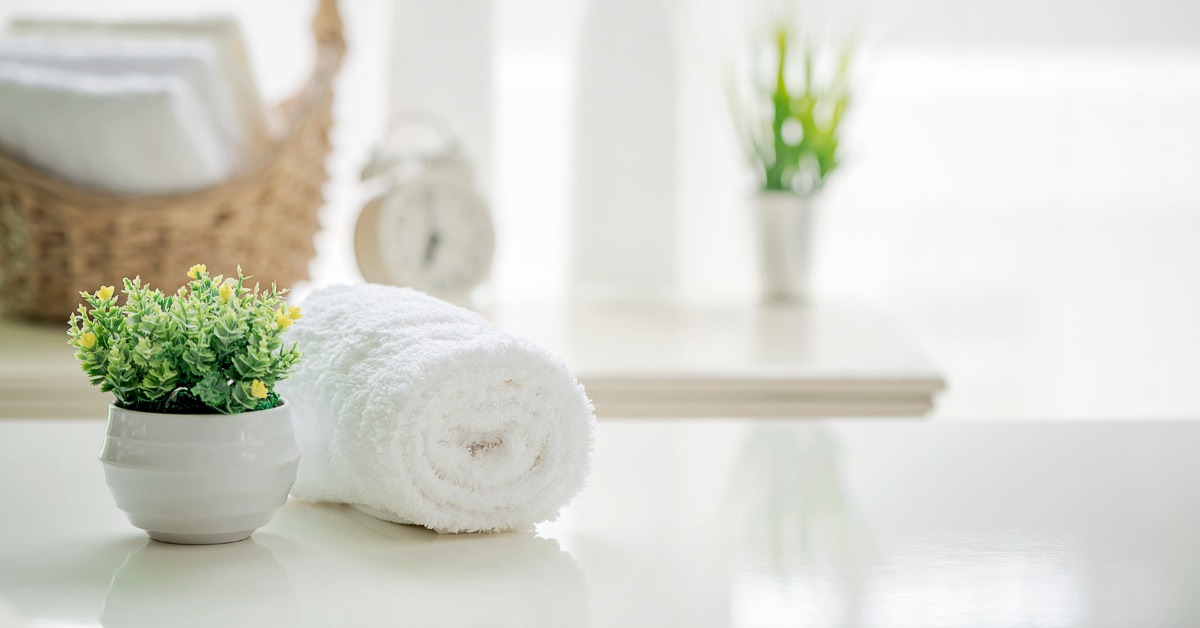 A rolled white spa towel beside a small potted plant on a white countertop, with a basket, clock, and plant in the background.