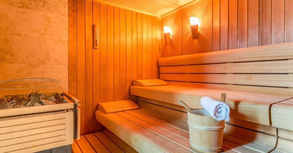 An indoor sauna with wooden bench seating, wall-mounted lights, a heater with stones, and a bucket with a ladle and towel on the bench.