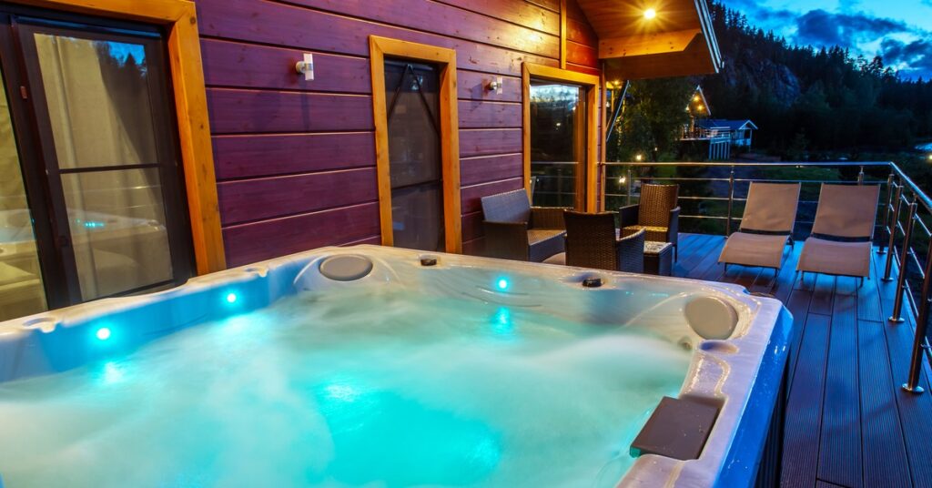 A hot tub with glowing blue water on a wooden deck beside a modern home at dusk, overlooking a scenic mountain landscape.