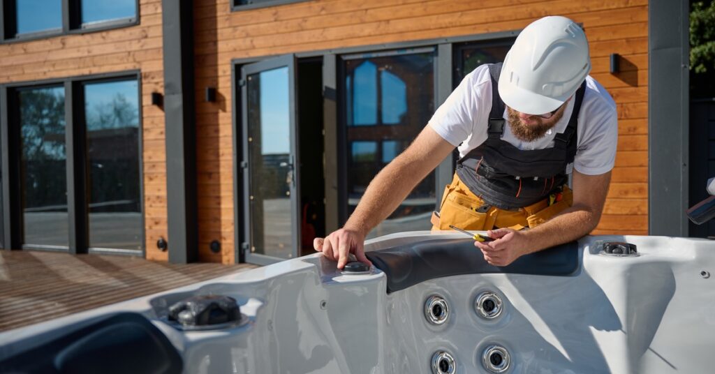 A technician wearing a white hard hat inspecting an empty hot tub on a backyard deck outside a modern wooden home.