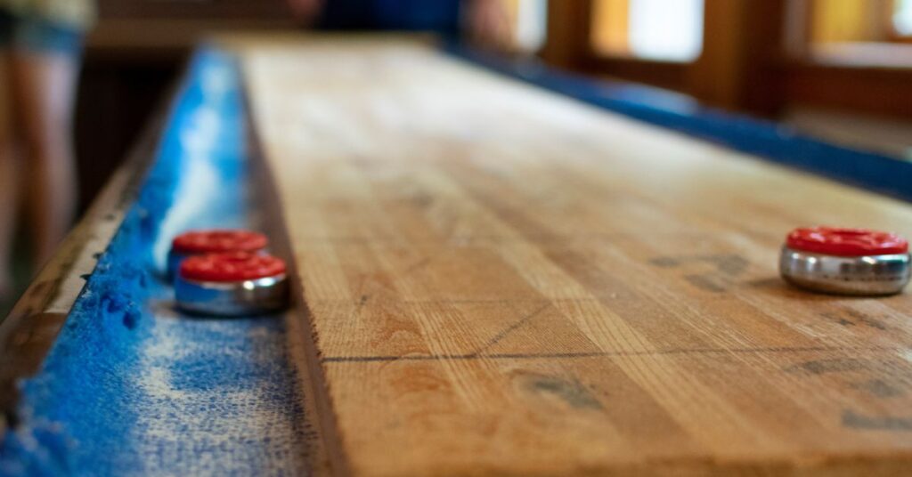 A shuffleboard table with red pucks resting on the polished wooden surface, with players blurred in the background.