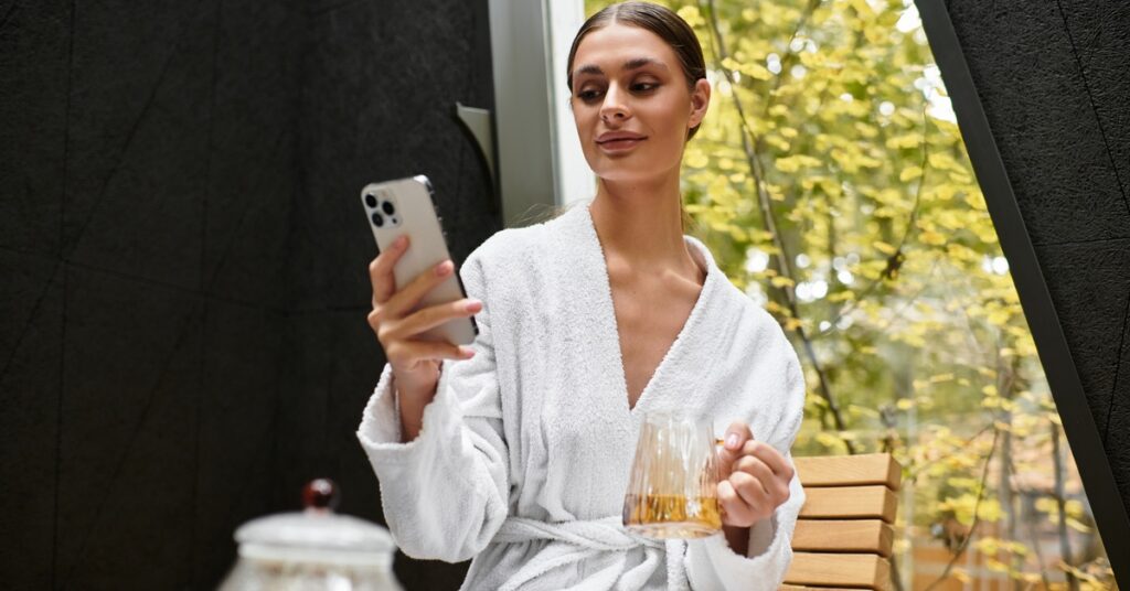 A young woman in a white robe relaxing in a sauna lounge area while holding a smartphone and a cup of tea.