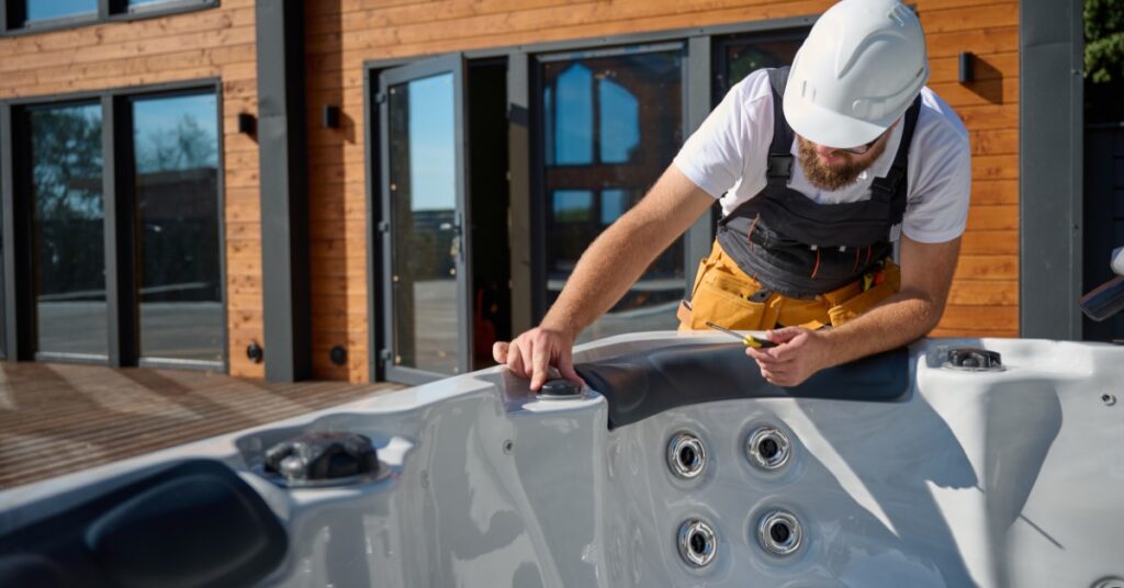 A technician servicing an outdoor hot tub outside a modern home with wood plank siding, checking the spa's controls and jets.