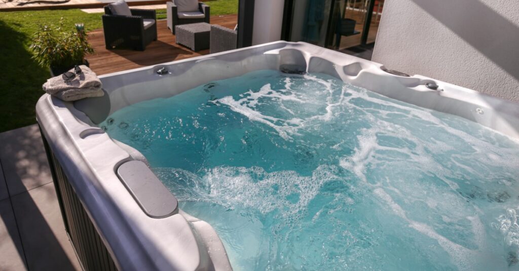 An outdoor hot tub with bubbling water on a backyard patio, overlooking a landscaped yard with a swimming pool and an outdoor seating area.