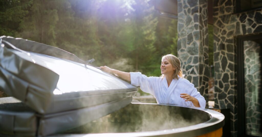 A woman in a white robe opens the cover of a steaming outdoor hot tub beside a stone-clad home.