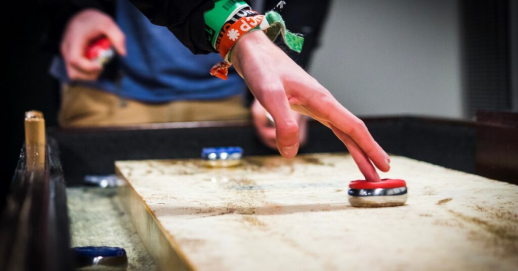 A person releasing a red shuffleboard puck with two fingers on a wooden shuffleboard table.
